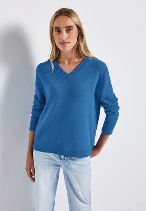Blauwe v-halstrui met ribbelaccenten, lange mouwen en een relaxte pasvorm. Gecombineerd met lichtblauwe denim jeans.