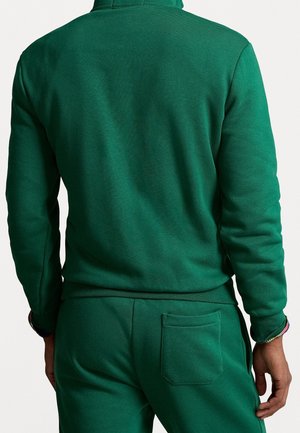 Mann trägt grünen Kapuzenpullover mit langen Ärmeln und passende Hose, von hinten vor einem einfarbigen hellen Hintergrund gezeigt.