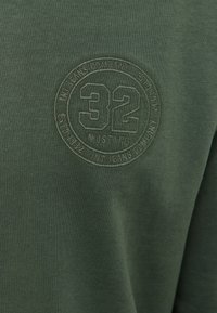 Žalias megztinis su raukšlių tekstūra, su apvaliu reljefiniu logotipu, kuriame centre yra "32" ir "MUSTANG".