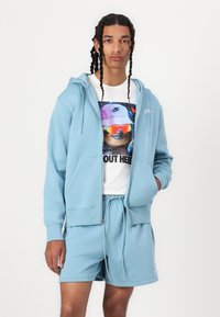 Lyseblå zip-up hettegenser med forkortte lommer over en grafisk hvit t-skjorte. Koordinerende lyseblå shorts med snor i livet, stoff synlig.