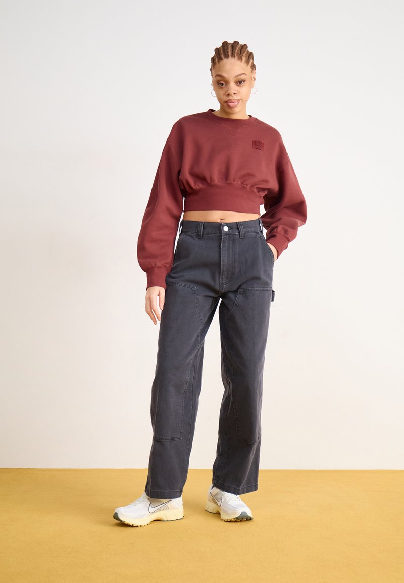 Bordeaux cropped sweatshirt, relaxte fit, gecombineerd met donkergrijze wijde broek met zakken en witte sportschoenen op een gele vloer.