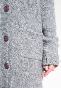 Grå fleecejacka med en texturerad yta, med runda bruna knappar och sidofickor. Ärmarna har en mjuk, lös passform.