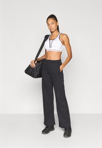 Biały sportowy biustonosz z czarnymi akcentami, luźne czarne spodnie dresowe i czarne buty sportowe. Modelka pozuje z czarną torbą podróżną.