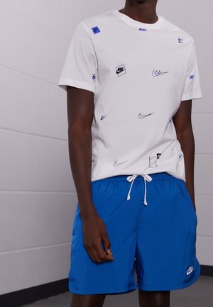 Persoon die een wit Nike T-shirt met meerdere logo's en blauwe Nike shorts met een witte trekkoord draagt, staand binnen tegen een lichte muur.