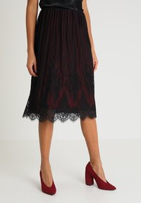 Gonna plissettata lunga fino al ginocchio di colore rosso scuro con orlo in pizzo. Abbinata a scarpe con tacco rosso. Texture morbida con una leggera lucentezza.