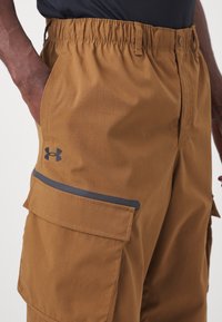 Hnědé cargo šortky vyrobené z lehké tkaniny, s elastickým pasem, bočními kapsami a černým logem jako akcentem.