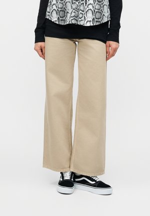 Személy bézs színű, bő szárú nadrágot és fekete-fehér tornacipőt visel, fehér háttér előtt áll, karjai lazán oldalra engedve.