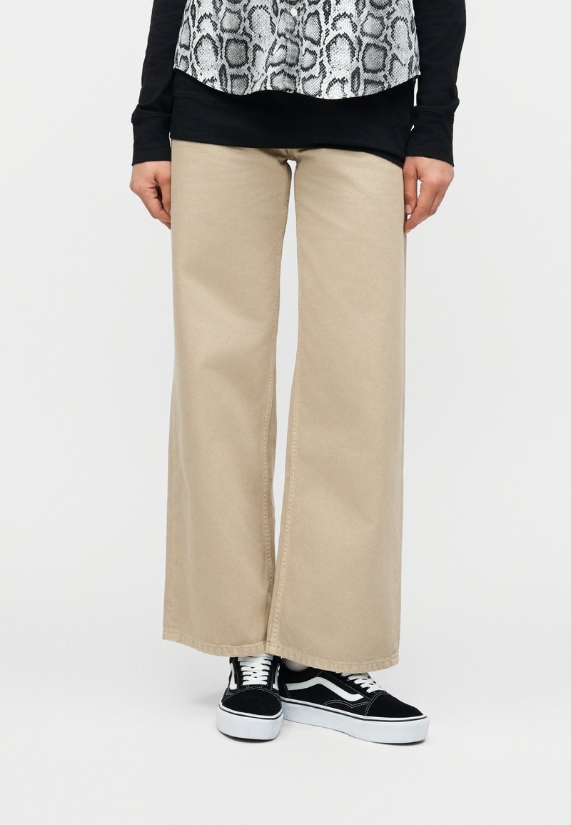 Άτομο που φοράει μπεζ παντελόνι με φαρδιά μπατζάκια και μαύρα-άσπρα αθλητικά παπούτσια, στεκόμενο με τα χέρια χαλαρά στα πλευρά, μπροστά σε λευκό φόντο.