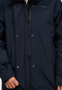 Giacca impermeabile blu realizzata in tessuto leggero, dotata di zip completa, bottoni a pressione e un colletto alto per una protezione aggiuntiva.