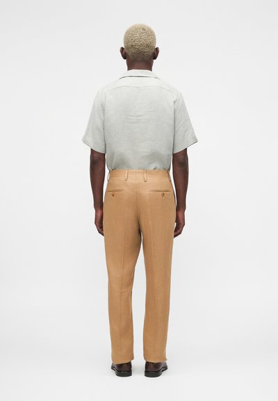 Άνδρας με κοντά ξανθά μαλλιά φοράει ανοιχτό γκρι κοντομάνικο πουκάμισο μέσα σε μπεζ παντελόνια, στέκεται με την πλάτη προς τα εμπρός σε λευκό φόντο.