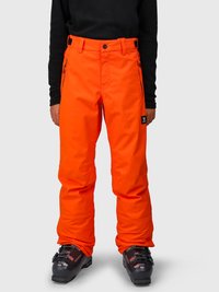 Person iført lyse orange skibukser med sorte skistøvler og en sort langærmet top, stående mod en plain baggrund.