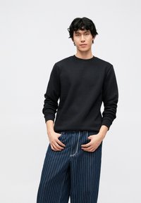 Fekete pulóver hosszú ujjakkal és kerek nyakkivágással, széles, csíkozott farmerral párosítva, kék-fehér mintákkal.