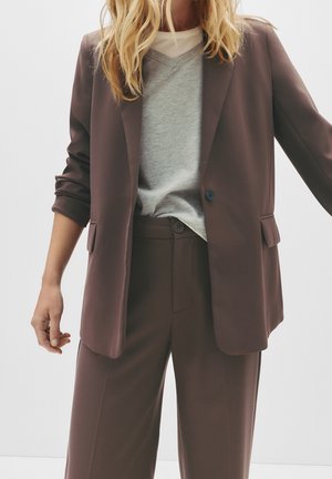 Femme portant un blazer marron et un pantalon assorti par-dessus un pull gris en V et un sous-vêtement blanc, debout devant un fond clair uni.