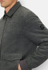Graue Wolljacke mit einem Reißverschluss, Kragen und zwei Fronttaschen. Mit einem strukturierten Stoff und einem Logo-Patch am linken Ärmel.