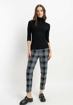 Schwarzes Rollkragenoberteil mit Dreiviertelärmeln, kombiniert mit blauen und schwarzen karierten Hosen. Das Model trägt schwarze Plateauschuhe.