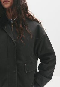 Veste noire avec fermeture éclair à l'avant, grand col et poches latérales ; présente un tissu texturé et des détails de couture visibles.