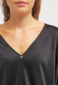 Black V-neck top made of smooth fabric, featuring a subtle triangular metal accent at the neckline and a relaxed fit.