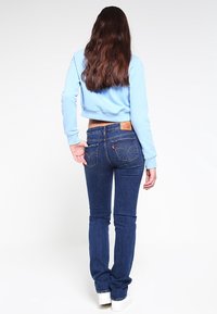 Sudadera recortada azul combinada con jeans ajustados azul oscuro que cuentan con bolsillos traseros y etiqueta de la marca en la cinturilla. Tenis blancos visibles.