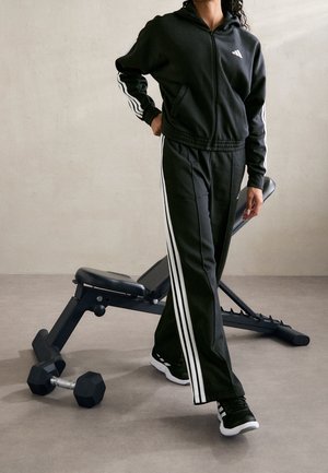 Person trägt schwarzen und weißen gestreiften Trainingsanzug und steht neben einer Trainingsbank und einer Hantel auf einem grauen Boden.