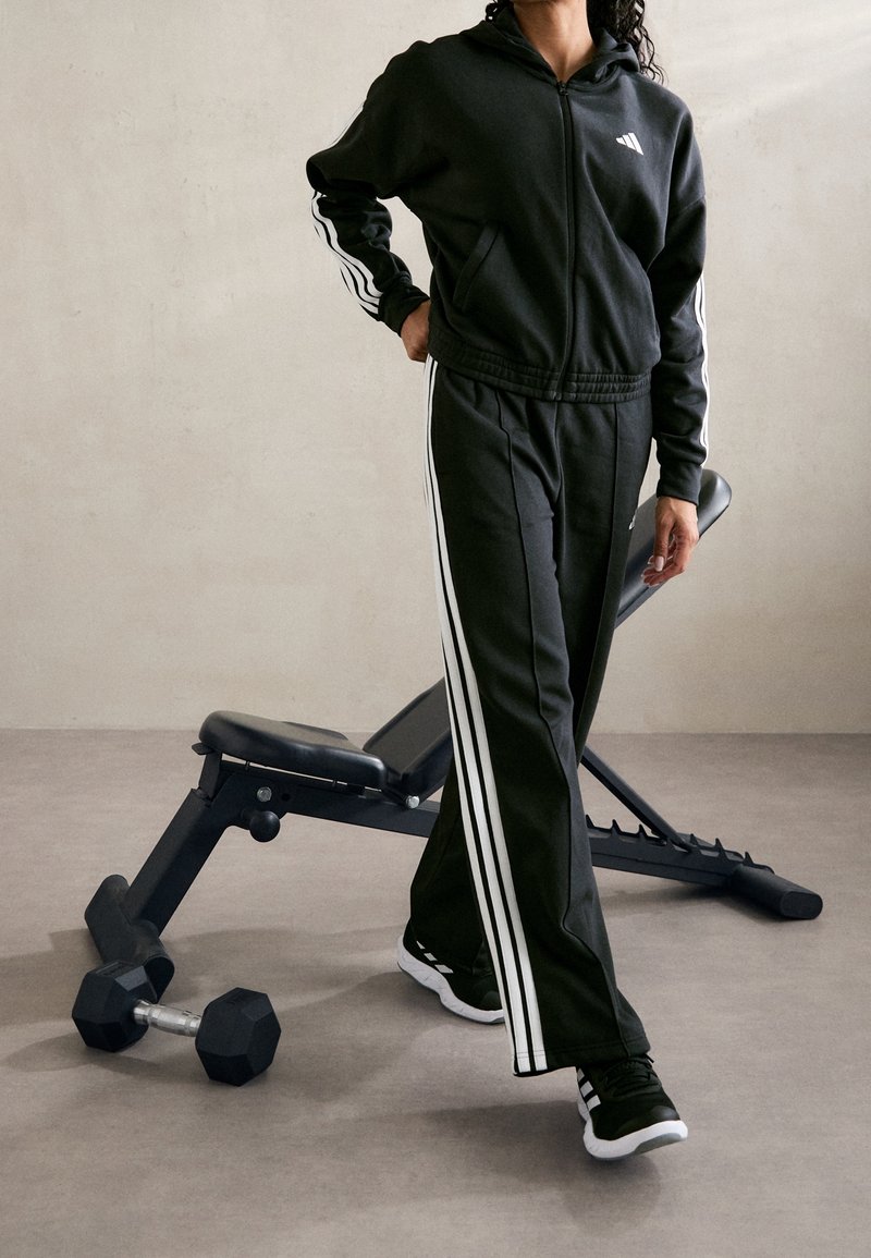 Osoba ubrana w czarno-biały paskowany dres stojąca obok ławki do ćwiczeń i hantla na szarej podłodze.