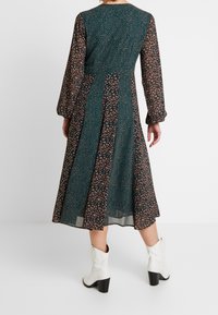 Blumenbedrucktes Kleid mit langen Ärmeln, grünem und schwarzem Stoff, leichtem Material und fließender Silhouette. Kombiniert mit weißen Absatzstiefeln.