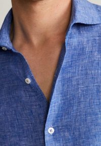 Camisa de lino azul con apariencia texturizada, que presenta un cuello y cierre frontal con botones. Los botones destacados son de color claro con un diseño sutil.