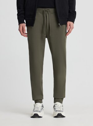 Uomo con pantaloni da jogging verde oliva con coulisse in vita, abbinati a scarpe da ginnastica bianche e grigie e una giacca nera con zip.