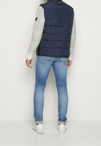 Marineblauer Puffer-Weste mit horizontalen Nähten, über einem hellgrauen Pullover getragen, kombiniert mit hellblauen Jeans und weißen Sneakers.