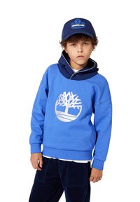 Gutt som har på seg en blå Timberland-cap, blå hettegenser med hvit tre-logo, hvit skjorte under, og mørkeblå kordfløyelsbukser.