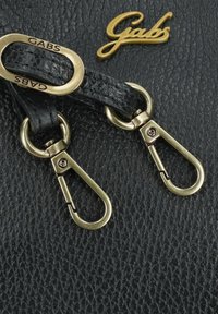Bolso de cuero negro texturizado que presenta herrajes en tono dorado, dos clips de metal y un prominente logotipo en relieve que dice "Gabs".