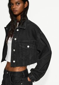 Fekete, rövid fazonú farmerdzseki klasszikus gallérral, ezüst gombokkal és elülső zsebekkel; rojtos szegélyével és széles ujjaival rendelkezik.