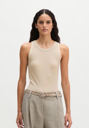 Femme aux longs cheveux foncés portant un haut beige sans manches rentré dans un pantalon gris taille haute avec une ceinture assortie sur fond blanc.