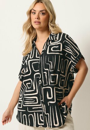 Blonde Frau, die ein kurzärmeliges schwarzes Hemd mit beigen, abstrakten labyrinthähnlichen Mustern trägt und mit einer Hand in der Tasche steht.