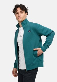 Giacca con zip teal, colletto alto, tasche laterali e patch logo sul petto. Indossata sopra una camicia bianca, con una texture liscia e una vestibilità comoda.