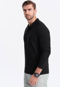 Musta pitkähihanen pikeepaita, jossa on kaulus ja slim fit -leikkaus. Yhdistetty vaaleanvärisiin housuihin ja mustaan kelloon ranteessa.