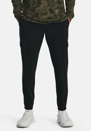 Mann trägt schwarze Cargojogger, olivgrünes langärmeliges Shirt und weiße Turnschuhe, steht mit den Händen in den Taschen vor weißem Hintergrund.