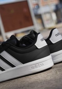 Baskets Adidas noir et blanc avec trois bandes blanches et logo Adidas sur le talon et la semelle, posées sur une surface en béton.