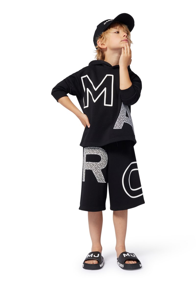 Felpa nera con lettere bianche sovradimensionate "M" e "A", pantaloni corti neri con "R" bianca e motivo testurizzato, cappellino nero abbinato e ciabatte nere.