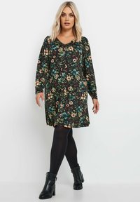 Pitkähihainen musta kukkamekko, jossa on värikkäitä kukkia, V-pääntie ja keräyksiä olkapäillä, yhdistettynä mustiin sukkahousuihin ja nilkkureihin.