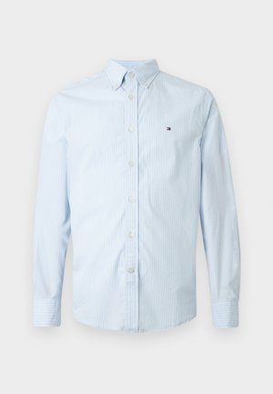 Lyseblå langærmet skjorte med knaplukning, der har diskrete hvide striber og et lille broderet logo på venstre bryst.