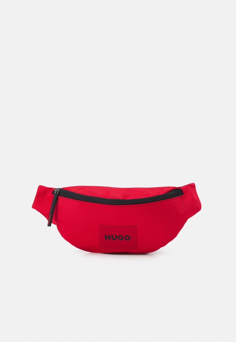 Banane en tissu rouge avec fermeture éclair noire et écusson logo "HUGO" à l'avant, présentée sur un fond clair uni.