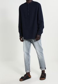 Camicia a maniche lunghe blu navy con colletto alla cinese e patta dei bottoni, abbinata a jeans azzurri risvoltati e sandali neri.