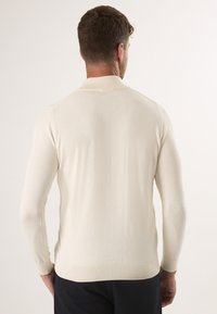 Cremfarbener gestrickter Pullover mit geripptem Kragen und Bündchen. Nahtdetails an den Schultern, taillierte Passform und glatte Textur.