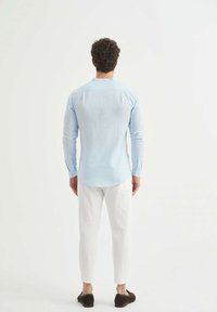 Chemise longue à manches longues bleu clair avec un col boutonné, associée à un pantalon blanc et des chaussures foncées, vue de dos.