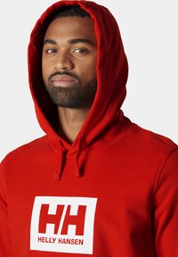 Sudadera roja con un logotipo blanco en el pecho. Cuenta con un bolsillo tipo canguro, capucha con cordón ajustable y puños de canalé. Fabricada con un tejido suave.