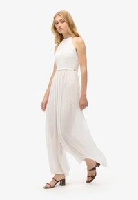 Abito bianco con scollo a halter, caratterizzato da un corpetto texturizzato, una gonna plissettata e un tessuto leggero e arioso. Tacchi a striscia marroni completano il look.