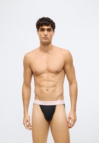 Jonge man met kort krullend haar, gekleed in zwart en roze Calvin Klein ondergoed, staande tegen een eenvoudige witte achtergrond.