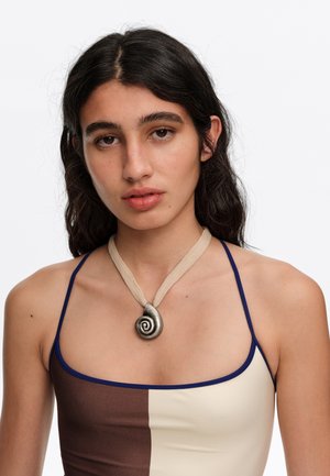 Joven con cabello largo y oscuro que lleva una camiseta sin mangas bicolor marrón y crema y un collar con colgante en espiral sobre cordón beige.