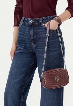 Mujer con vaqueros azul oscuro y una blusa burdeos con textura acanalada, sosteniendo un pequeño bolso marrón acolchado con una correa de cadena plateada.