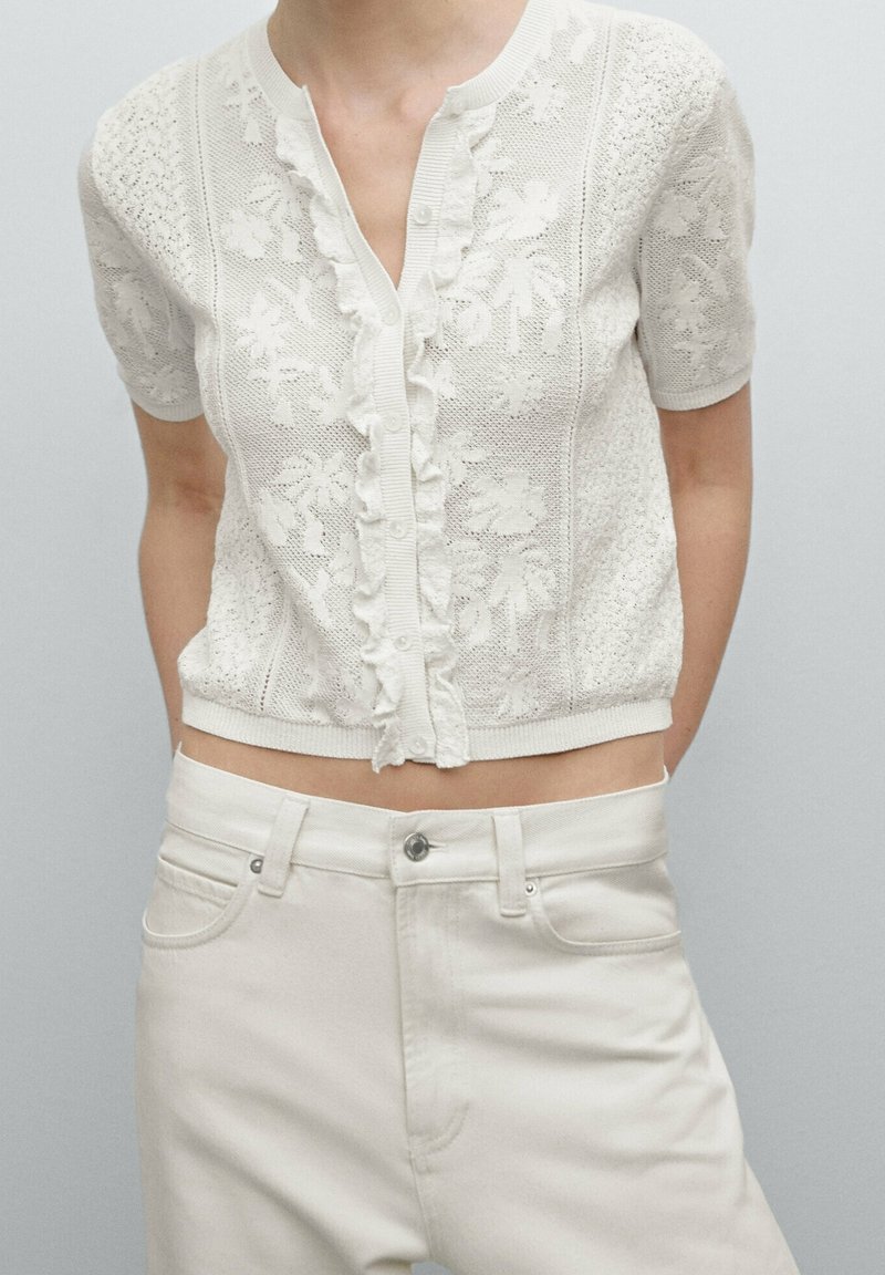 Femme portant un cardigan tricoté blanc à manches courtes avec motif floral et bordure volantée, assorti à un pantalon blanc cassé taille haute.